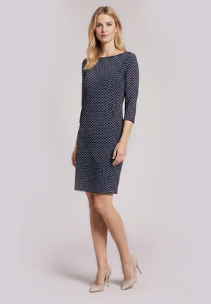 Frau steht, trägt ein figurbetontes knielanges marineblaues gemustertes Kleid mit dreiviertel Ärmeln und beigen High Heels vor hellem Hintergrund.