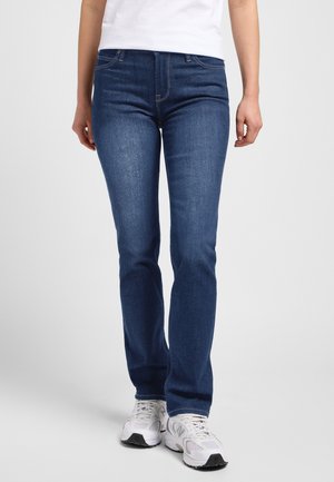 Osoba nosząca ciemnoniebieskie dopasowane dżinsy i białe trampki, stojąca z luźno opuszczonymi rękami na tle o neutralnym kolorze.