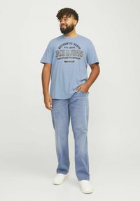 Camiseta de manga corta de color azul claro con texto gráfico, combinada con jeans azules claros y zapatillas blancas, llevada por un hombre con cabello rizado.