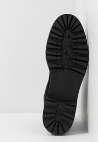 Semelle en caoutchouc noir avec un motif de texture, comportant des formes en losange et des lignes. Bords arrondis, usée à un angle contre un fond uni.