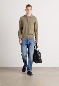 Oliven grønn langermet polo-skjorte, blå denim jeans, svart bærebag og svarte sko. Modellen står mot en ensfarget bakgrunn.