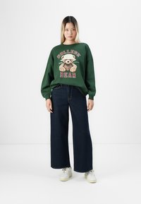 Una giovane donna indossa un felpa verde con un orsacchiotto bendato e il testo "College Bear", jeans larghi scuri e sneaker bianche.