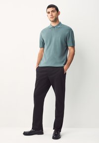 Polohemd in sanftem Türkis, kurzärmlig, mit Reißverschluss. Eng geschnittene schwarze Hose und glänzende schwarze Schuhe vervollständigen das Outfit. Einfarbiger Hintergrund.