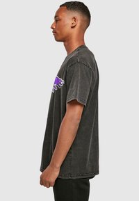 Černé, oversized bavlněné tričko s krátkými rukávy, zdobené fialovým grafickým potiskem na levém rameni, vystavené na modelu z boku.