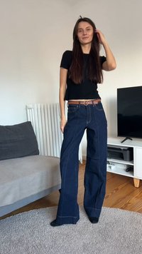 Mörkblå jeans med vida ben och synliga sömmar, använda med ett brunt bälte med ett runt spänne. Platt textur, bomullsdenimtyg.