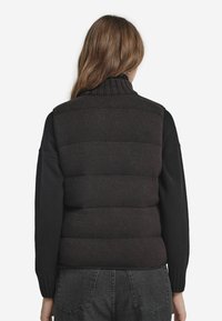 Svart stickad väst med hög krage, med en puffig nederdel och ribbade detaljer på ärmarna, bärs över mörka byxor.