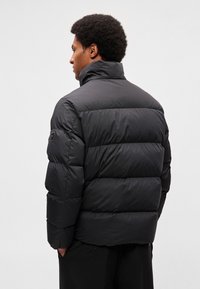 Veste matelassée noire avec un col montant, un motif matelassé horizontal et des poignets élastiques. Le tissu a une texture lisse et résistante à l'eau.