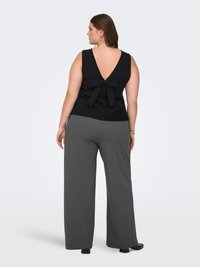 Vrouw met mouwloze zwarte top met strikdetail aan de achterkant en grijze broek met wijde pijpen, staand met haar rug naar de camera op een lichte achtergrond.