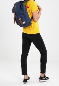 Granatowy plecak z brązowymi skórzanymi akcentami, duża klapa i przednia kieszeń; noszony z żółtą koszulą i czarnymi obcisłymi dżinsami.