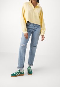 Keltainen, puolipitkä vetoketjullinen pitkähiha-pulloveri logolla; vaaleansiniset, korkeavyötäröiset farkut, joissa kulutettuja yksityiskohtia; vihreät mokkanahkaiset lenkkarit.