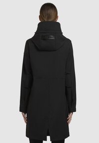 Parka imperméable noire avec un col haut, des coutures visibles et un patch logo dans le dos. Présente un design de longueur intermédiaire et des fentes sur les côtés.