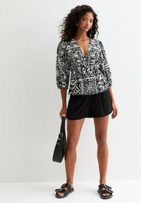 Blouse à motifs noir et blanc avec un col en V, des manches 3/4 et un design peplum, associée à un short noir et un sac à main noir.
