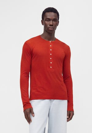 Homme portant un t-shirt henley orange brûlé à manches longues avec des boutons blancs et un pantalon gris clair, debout devant un fond clair uni.
