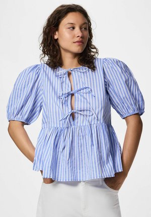 Femme portant un chemisier à rayures verticales bleu et blanc avec des manches bouffantes et trois nœuds noués sur le devant, associé à un pantalon blanc.