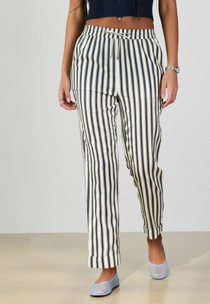 Kvinde iført sort-hvide bukser med lodrette striber, sort kort top og lyseblå mesh slip-on sko, stående på beige gulv.