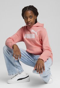 Felpa rosa con logo "PUMA" bianco, abbinata a jeans blu chiaro e sneakers bianche con dettagli neri. Texture del tessuto liscia.