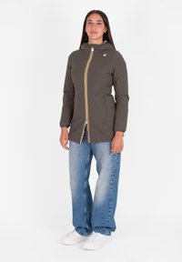 Olijfgroene pufferjack met een capuchon, voorkant met ritssluiting met gele accenten en zijzakken, gedragen over losse blauwe jeans.