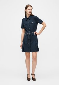 Γυναίκα που φοράει φόρεμα μπροστινής κούμπωμα σε σκούρο μπλε με κοντά μανίκια, αλυσίδα μέσης και μαύρα παπούτσια Mary Jane, στέκεται με το ένα χέρι στη μέση.