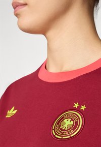 Maillot bordeaux avec un col rose, orné d'un logo jaune brodé de la Deutscher Fußball-Bund et de trois étoiles au-dessus.