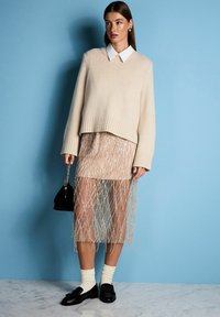 Beiger Strickpullover mit geripptem Saum, darüber ein transparenter, hautfarbener Rock mit silbernen Perlenfransen. Schwarze Handtasche und Schuhe vervollständigen den Look.