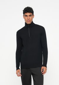 Schwarzer Zip-Kragen-Pullover mit langen Ärmeln, strukturiertem Stoff und taillierter Passform, kombiniert mit dunklen Hosen, vor einem weißen Hintergrund präsentiert.