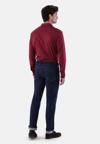 Langärmliges burgunderfarbenes Hemd mit Kragen, kombiniert mit dunkelblauen Jeans mit umgeschlagenen Säumen. Braune Schuhe ergänzen das Outfit.