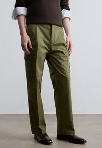 Olivzaļas krāsas cargo bikses ar brīvu piegriezumu, ar sānu kabatām un gludu auduma faktūru. Kombinētas ar tumši brūnām kurpēm.