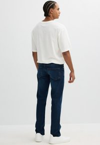 Camiseta de algodón blanca, ajuste relajado; combinada con jeans de mezclilla azul oscuro con bolsillos traseros. El modelo lleva zapatillas blancas y está de pie frente a un fondo claro.
