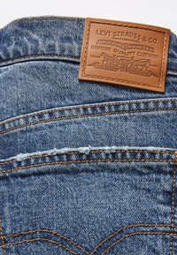 Jeans de denim azul con un parche de cuero marrón claro con marca en relieve, costuras naranjas en contraste y detalles en el bolsillo trasero.