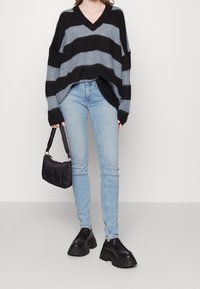 Persona che indossa un maglione oversize a righe nere e grigie, jeans skinny blu chiaro, scarpe chunky nere e tiene in mano una piccola borsa nera.
