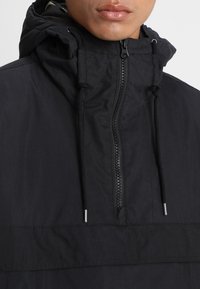 Svart pullover-jacka med hög krage, justerbara snoddar och central dragkedja. Tillverkad av ett lätt, texturerat tyg.