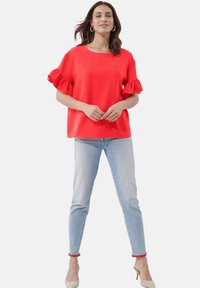 Rotes kurzärmliges Oberteil mit Rüschendetails, kombiniert mit hellblauen Skinny-Jeans. Die Jeans besitzen einen roten Pom-Pom-Besatz am Knöchel.