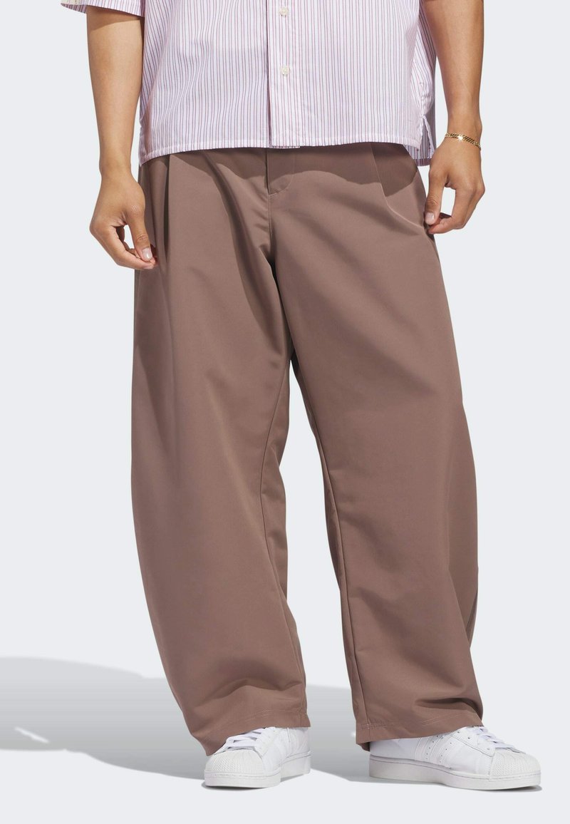Person wearing loose brown pants, white sneakers, and a short-sleeve pink and white striped shirt, standing against a plain background.