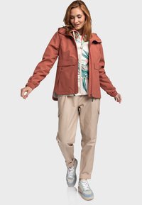 Veste rouille imperméable avec capuche, deux poches avant et poignets élastiques, portée sur un haut floral blanc et un pantalon cargo beige.
