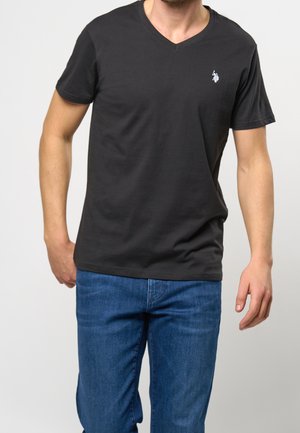 Mand iført sort T-shirt med V-hals og lille hvid logo på venstre bryst samt blå jeans, stående foran en ensfarvet lys baggrund.