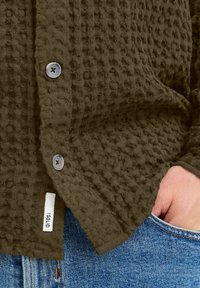 Camisa de botones marrón con textura y patrón de tejido de waffle, con botones oscuros y una pequeña etiqueta blanca, combinada con jeans de mezclilla.