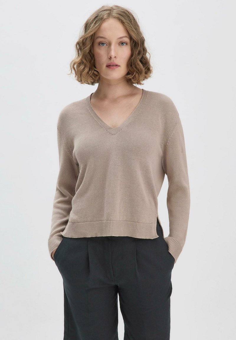 Beige V-Ausschnitt-Pullover mit langen Ärmeln, geripptem Saum und subtiler Textur, kombiniert mit dunkel gefärbten, hoch taillierten Hosen.