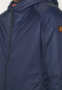 Giacca blu navale con cappuccio, zip frontale e un accento arancione. Il materiale sembra liscio e leggero, con una leggera lucentezza.