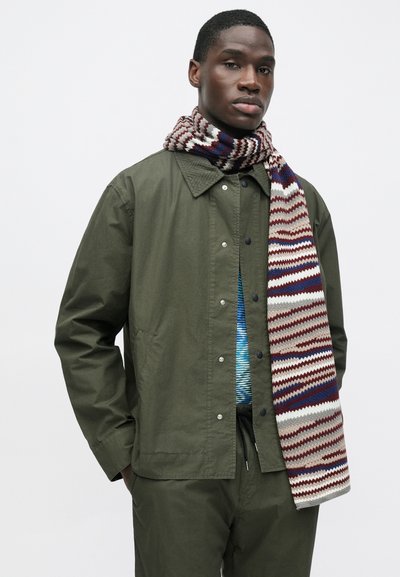 Olivovo zelená ľahká bunda s golierom a cvokmi, doplnená viacfarebným pruhovaným pleteným šálom v burgundských a sivých tónoch.