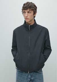 Leichte schwarze Jacke mit Reißverschluss vorne, Stehkragen und zwei Taschen vorne. Getragen mit hellblauen Jeans. Glatte Stoffstruktur.