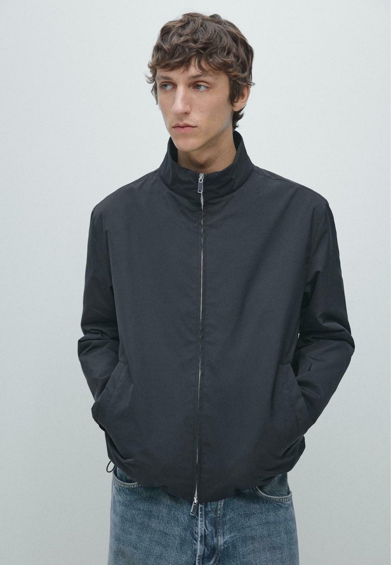 Leichte schwarze Jacke mit Reißverschluss vorne, Stehkragen und zwei Taschen vorne. Getragen mit hellblauen Jeans. Glatte Stoffstruktur.