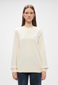 Κρέμα χρώματος φούτερ φτιαγμένο από μαλακό ύφασμα, με στρογγυλή λαιμόκοψη, ρίγες στους ώμους και μικρό τυπωμένο λογότυπο. Συνδυάζεται με σκούρα τζιν.