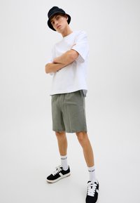 Camiseta blanca de manga corta, pantalones cortos oliva claros, sombrero tipo cubo negro, zapatillas negras con detalles blancos, y calcetines blancos con logo.