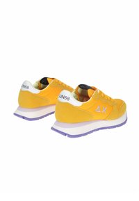 Geel-suède sneakers met een witte hielcounter, paarse zool en contrasterende zwart-witte branding op de tong en hiel.