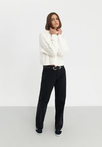 Pull cropped en tricot blanc, jean noir taille haute et baskets sombres. Le modèle se tient avec les mains près du cou, le regard légèrement tourné vers le haut.