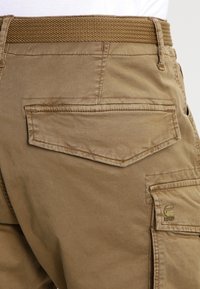 Pantaloni cargo marroni con una vita strutturata, caratterizzati da tasche posteriori e dettagli di cucitura, realizzati in tessuto resistente. Logo sul lato.