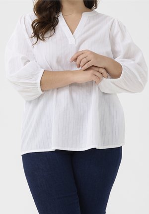 Kobieta z długimi włosami, ubrana w białą bluzkę z długim rękawem z delikatnymi pionowymi paskami oraz ciemnoniebieskie dżinsy, z rękami skrzyżowanymi na wysokości talii.