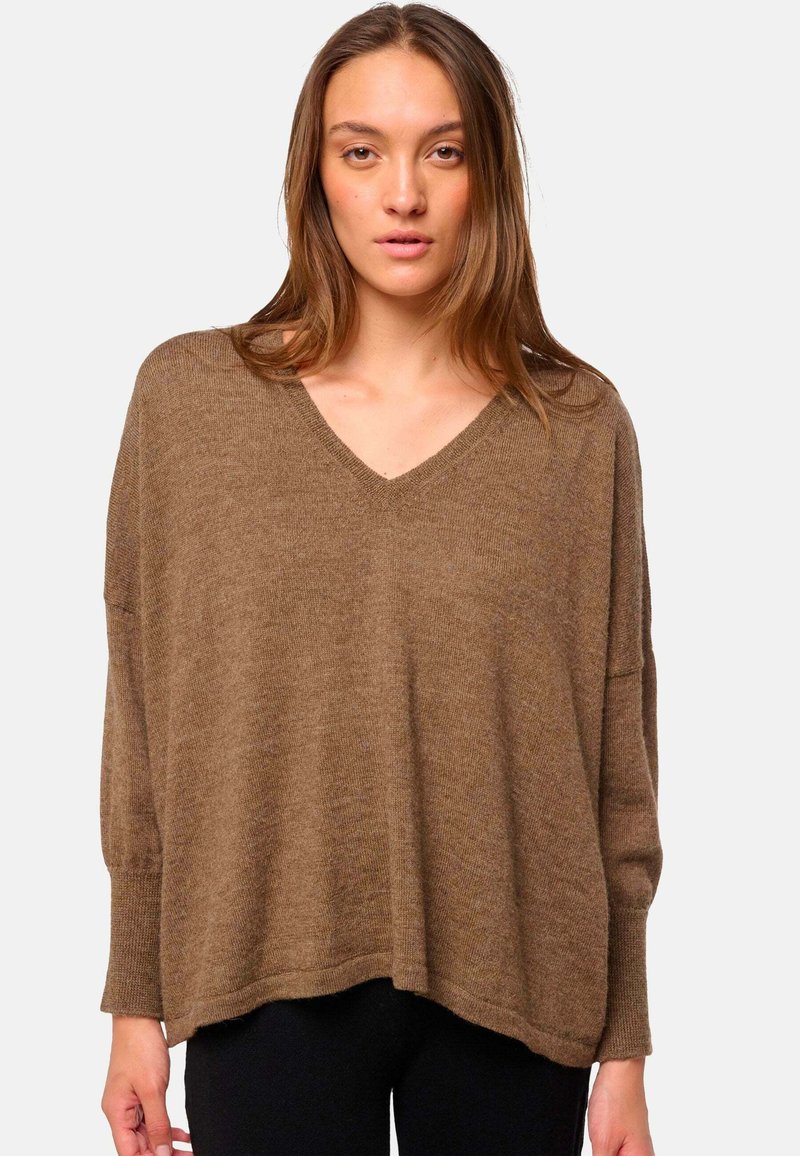 Jeune femme avec de longs cheveux bruns portant un pull marron ample à encolure en V et un pantalon noir, se tenant devant un fond clair.