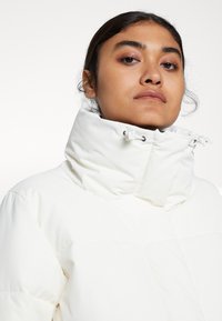 Doudoune blanche avec col haut et cordons de serrage ajustables ; texture lisse et motif en losanges à la surface.