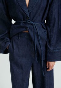 Denim obleka, ki vključuje temnordeč oversized jakno z velikimi gumbi, pasom na vezanje in ujemajoče se hlače z bočnimi žepi. Vidna teksturirana tkanina.